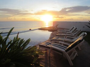 Une piscine à débordement avec des chaises longues donnant sur l’océan, au moment du coucher du soleil.