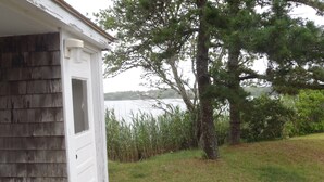 Pieni, yhden kerroksen rakennus, jonka ulkoseinät ovat valkoiset ja jossa on ovi. Se sijaitsee nurmikon vieressä, taustalla näkyy puu ja vesialue.