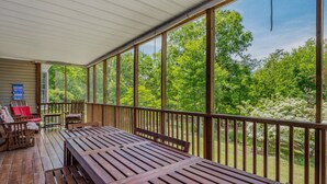 A covered outdoor deck with wooden furniture and a view of a forest. - Blue Badger Cottage-wifi, screen porch, fire pit, game room, king bed, fireplace (Berkeley Springs)