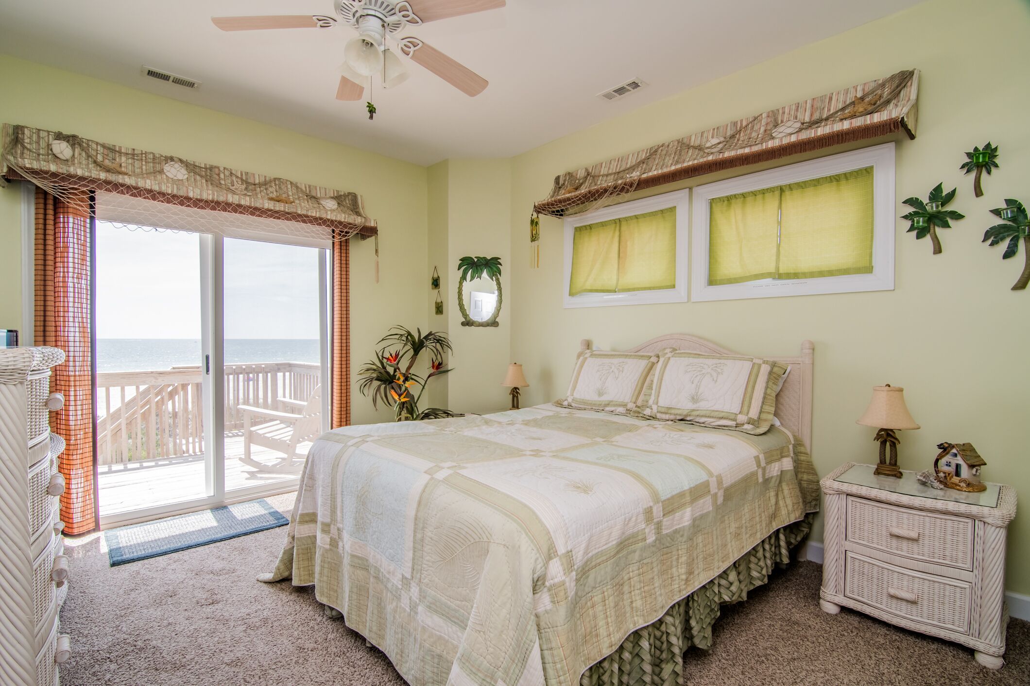 Une chambre à coucher comprenant un lit, une table de chevet, une commode, un ventilateur de plafond, une fenêtre avec des rideaux, une vue sur l’océan et un balcon.