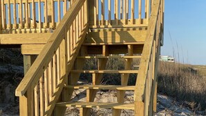 Un escalier en bois mène à une terrasse en bois dotée d’une rambarde, le tout étant situé sur un terrain sablonneux.
