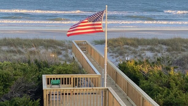 Une promenade en bois menant à une plage, avec un drapeau américain flottant au sommet d’un mât.