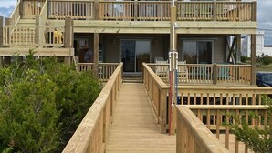 Une maison en bois à deux étages, dotée d’un balcon et d’une terrasse en bois menant à l’entrée, sous un ciel dégagé.
