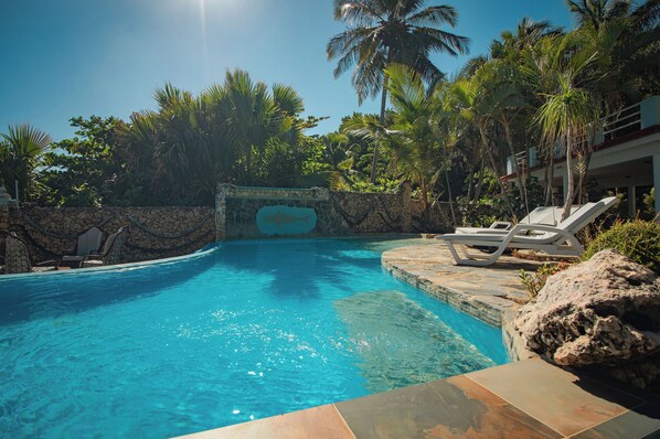 A clear blue pool surrounded by stone walls and palm trees, with lounge chairs and a stone pathway leading to the pool.