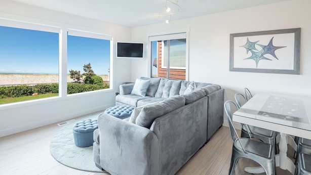 Een moderne woonkamer met een grijze bank, een glazen eettafel en uitzicht op het strand.