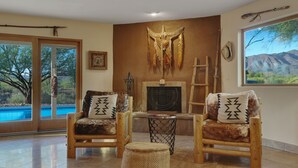 A room with two wooden chairs with patterned cushions, a fireplace, a window with a mountain view, and a pool visible through the glass doors. - Old Western Sonoran Desert Getaway (Cave Creek)
