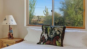 A bedroom with a large window, a wooden nightstand with a lamp, and a colorful pillow. - Old Western Sonoran Desert Getaway (Cave Creek)