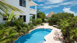 A pool area with a white building, a thatched-roof structure, and lush greenery.