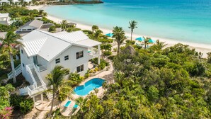 A large white house with a swimming pool, surrounded by lush greenery and a beach.