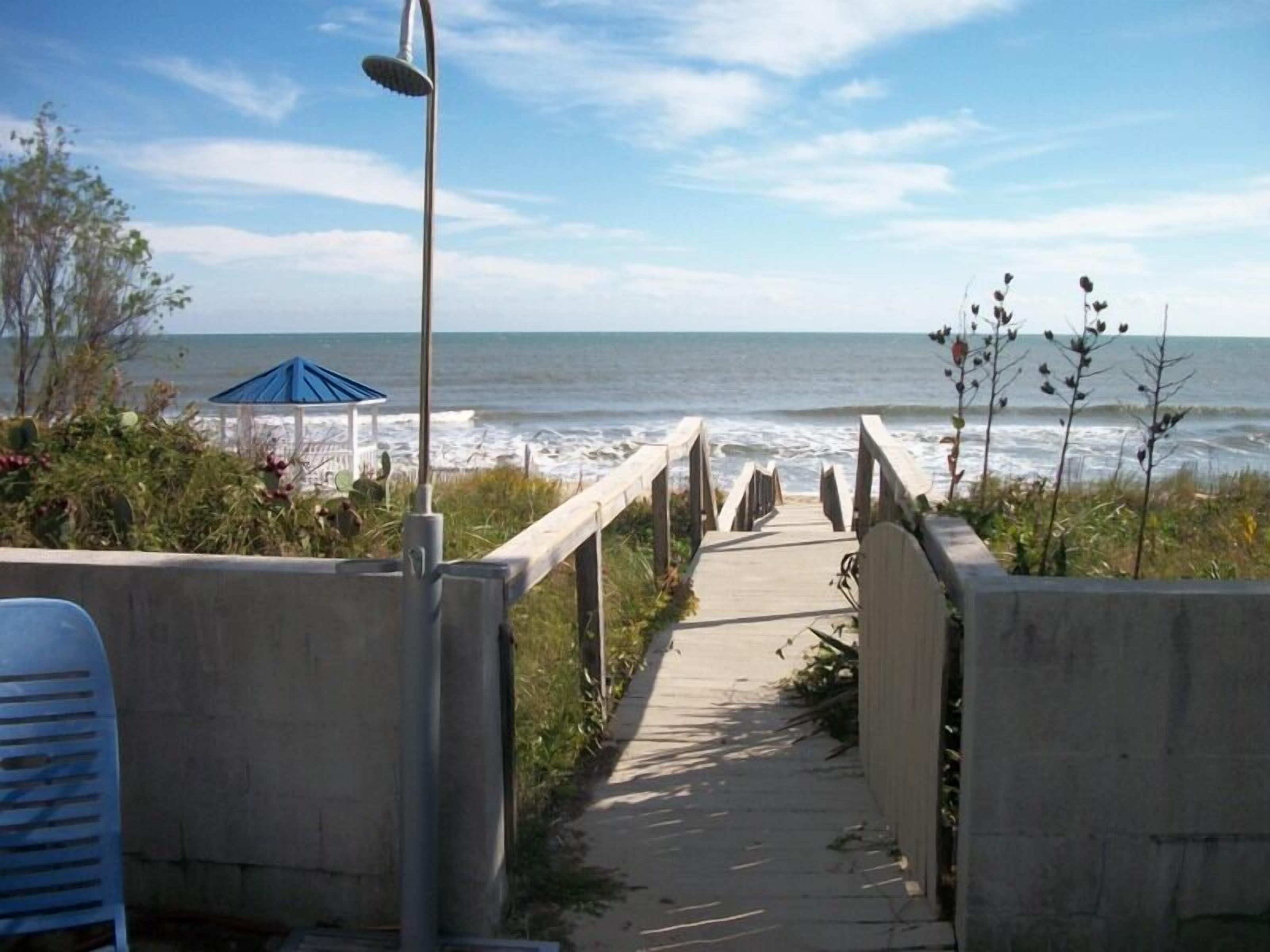 A concrete pathway leading to a beach with a blue umbrella and a metal lamp post.