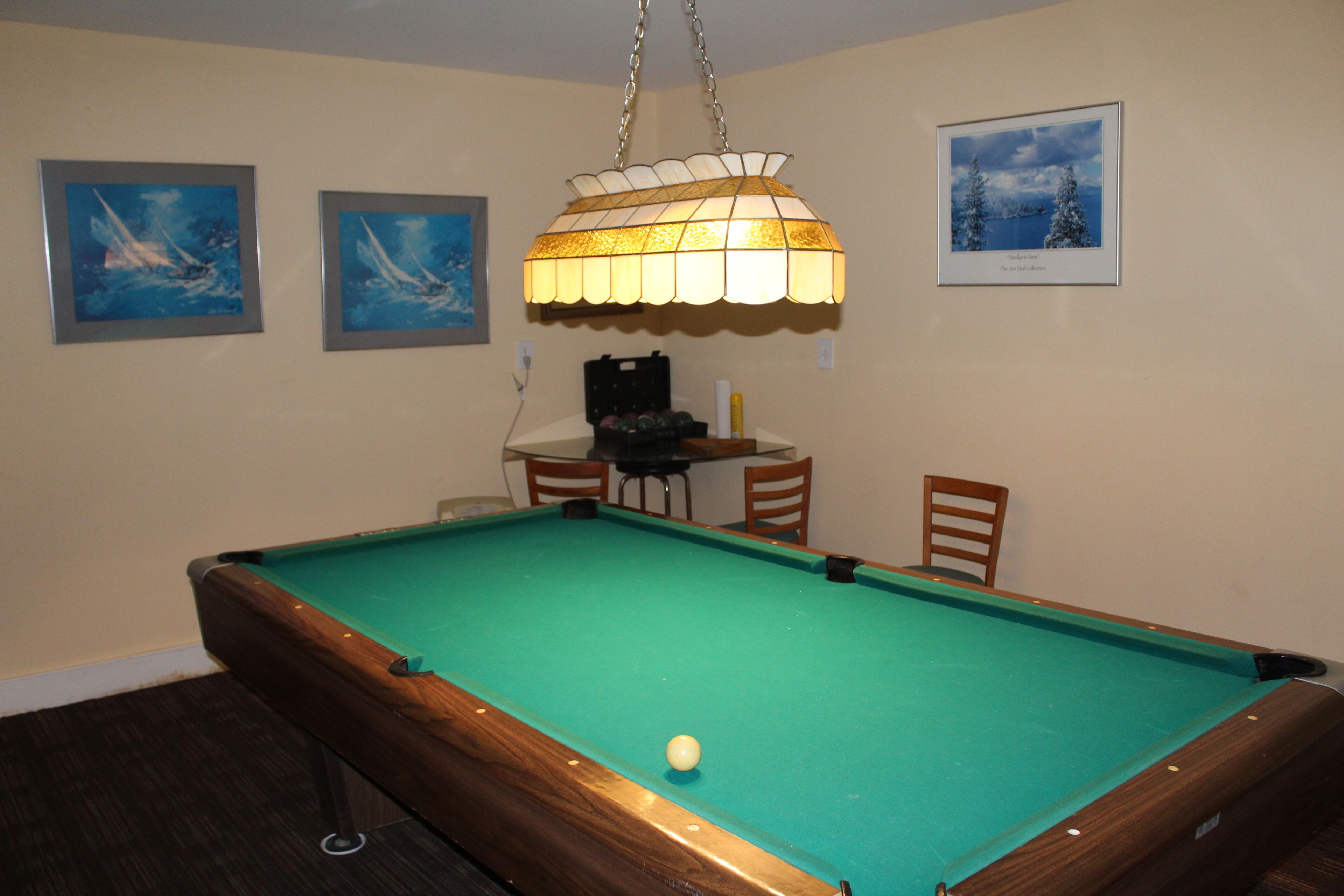 A pool table with a green felt surface, a hanging lamp, and two framed paintings on the wall.
