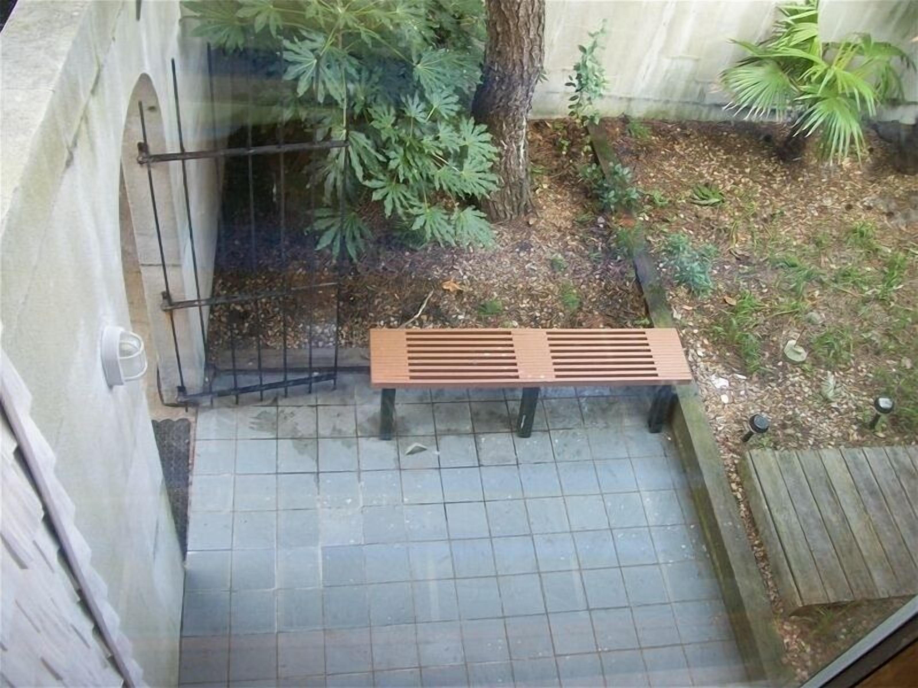 A wooden bench in a garden area with a tiled floor, surrounded by plants and a tree.