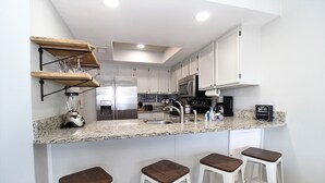 A modern kitchen with white cabinets, a granite countertop, a stainless steel refrigerator, a microwave, and a coffee machine. There are three bar stools with wooden seats. - Spacious Beachside Condo (Orange Beach)