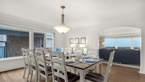 A dining area with a table set for four, white chairs, a chandelier, and a view through the window. - OCEAN FRONT: PACIFIC HAVEN:  On the Prom: Seaside Vacation Homes, Inc Managed  (Seaside)