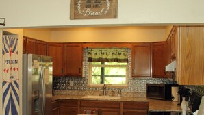 A kitchen with wooden cabinets, a stainless steel refrigerator, a granite countertop, and a window with a green curtain. - Come Live on the farm  (Starke)