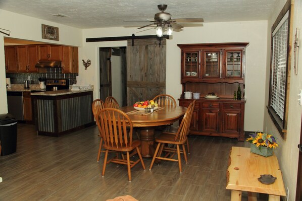 A kitchen with wooden cabinets, a dining table with chairs, a ceiling fan, and a small table with a vase of flowers. - Come Live on the farm (Starke)