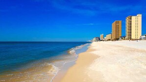 Uma praia com céu azul límpido, areal arenoso e edifícios altos ao fundo.
