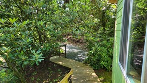 A wooden deck leading into a lush garden area with dense foliage and a small pond. - Pebble Creek Magical Retreat 2BR 1.5BA Shelter on Stream. Camp/Sleep 10/Linens
 (Laurel Fork)