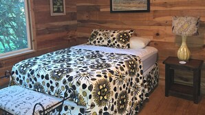 A wooden cabin bedroom with a bed, a nightstand, a window, and a framed picture on the wall. - Pebble Creek Magical Retreat 2BR 1.5BA Shelter on Stream. Camp/Sleep 10/Linens
 (Laurel Fork)