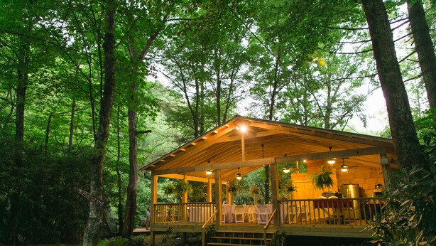 A wooden cabin with a porch, surrounded by trees. - Pebble Creek Magical Retreat 2BR 1.5BA Shelter on Stream. Camp/Sleep 10/Linens
 (Laurel Fork)