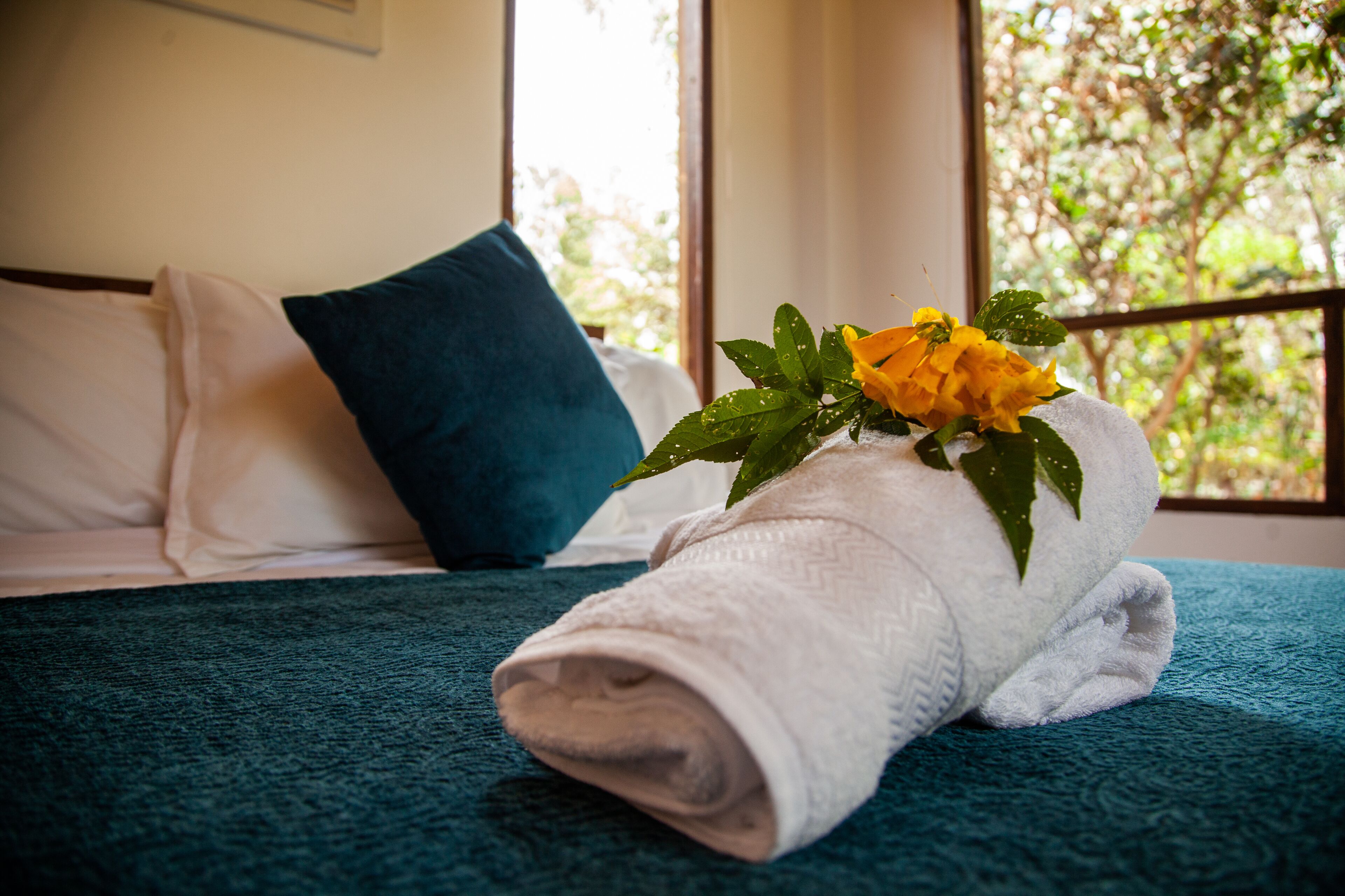 Ein ordentlich bezogenes Bett mit einer blauen Decke, einem weißen Kopfkissen und einem aufgerollten weißen Handtuch mit einer gelben Blume.