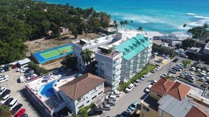 Un hotel de varios pisos con piscina en la azotea, junto a una playa y una cancha de tenis.