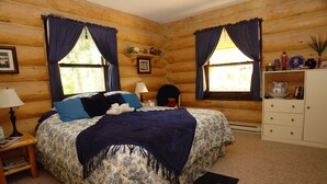 A log cabin bedroom with a bed, two windows with curtains, a nightstand, a dresser, and a small shelf with items. - Gray's room Garrison Inn Montana bed and breakfast (Kalispell)