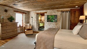 Un dormitorio con pared de piedra, techo de madera, cama con colcha, una silla, cómoda y un cuadro.