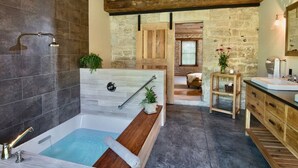 Un baño moderno con bañera, ducha, detalles de madera, paredes de piedra y vista a un dormitorio.