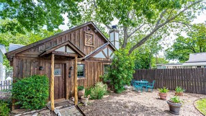 A rustic wooden cabin with a stone pathway leading to the door, surrounded by a fenced yard with potted plants and a blue metal table.