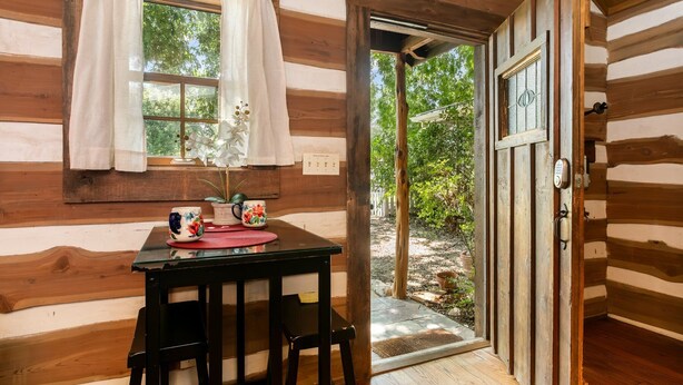 A wooden cabin interior with a dining table, chairs, a window with curtains, and a door leading outside.