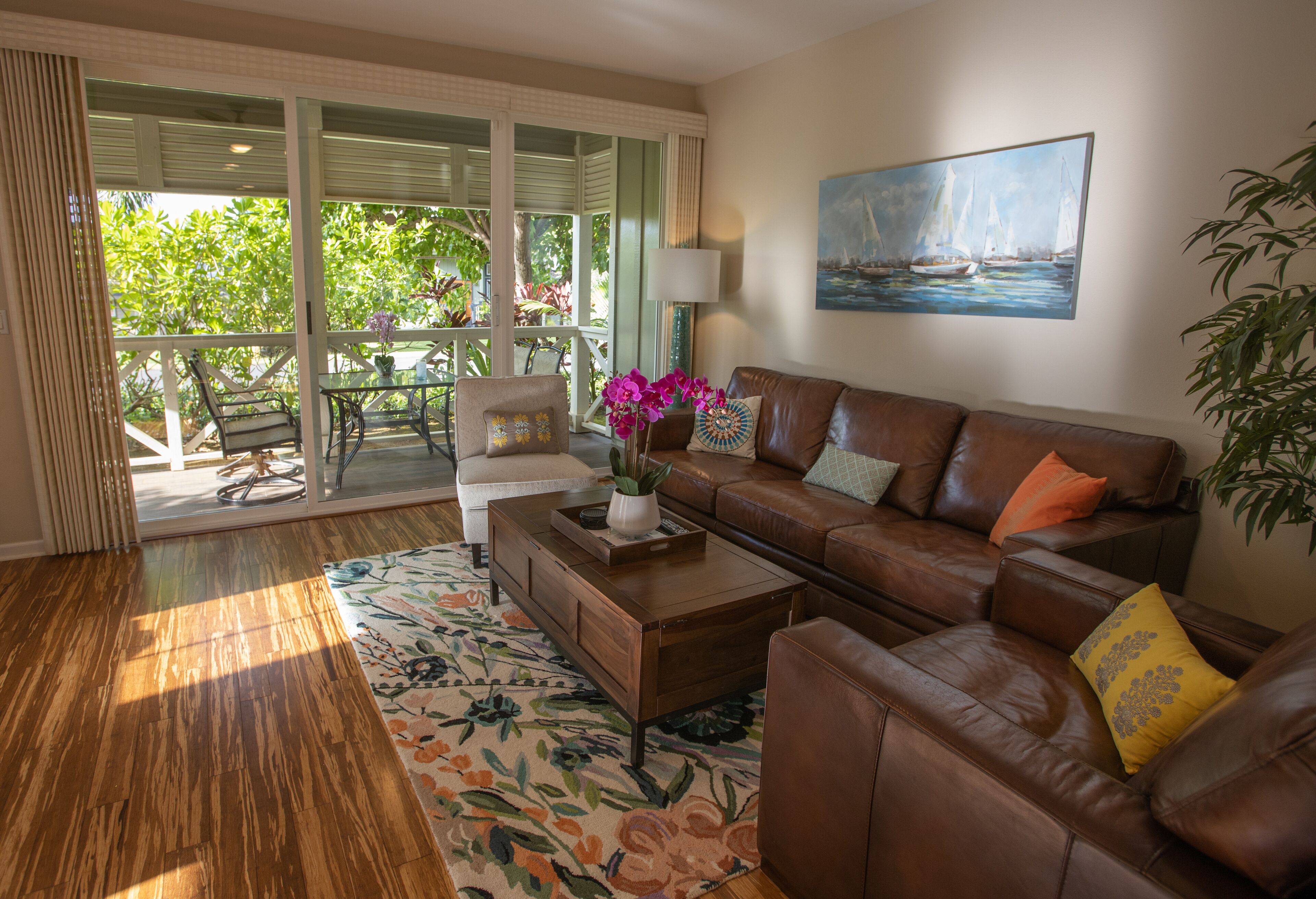 A living room with a brown leather sofa, a coffee table, and a floral rug. There is a painting of a sailboat on the wall and a potted plant on the right.