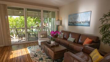 A living room with a brown leather sofa, a coffee table, and a floral rug. There is a painting of a sailboat on the wall and a potted plant on the right.
