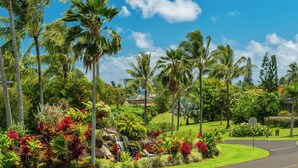 A lush tropical garden with palm trees and a waterfall, leading to a house with a red roof.