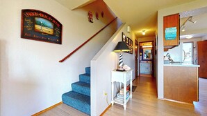 A staircase with a blue carpet leading to a landing with a lamp and a small table. A framed picture of a beach house is on the wall. - OCEAN FRONT: BEACHTOPIA South:  Seaside Vacation Homes, Inc Managed  (Seaside)