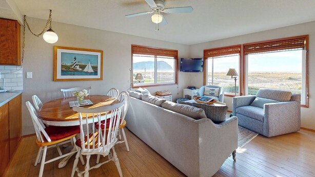 A spacious living area with a dining table, a sofa, and a couch. There are large windows with blinds, a ceiling fan, and a painting on the wall. - OCEAN FRONT: BEACHTOPIA South:  Seaside Vacation Homes, Inc Managed  (Seaside)