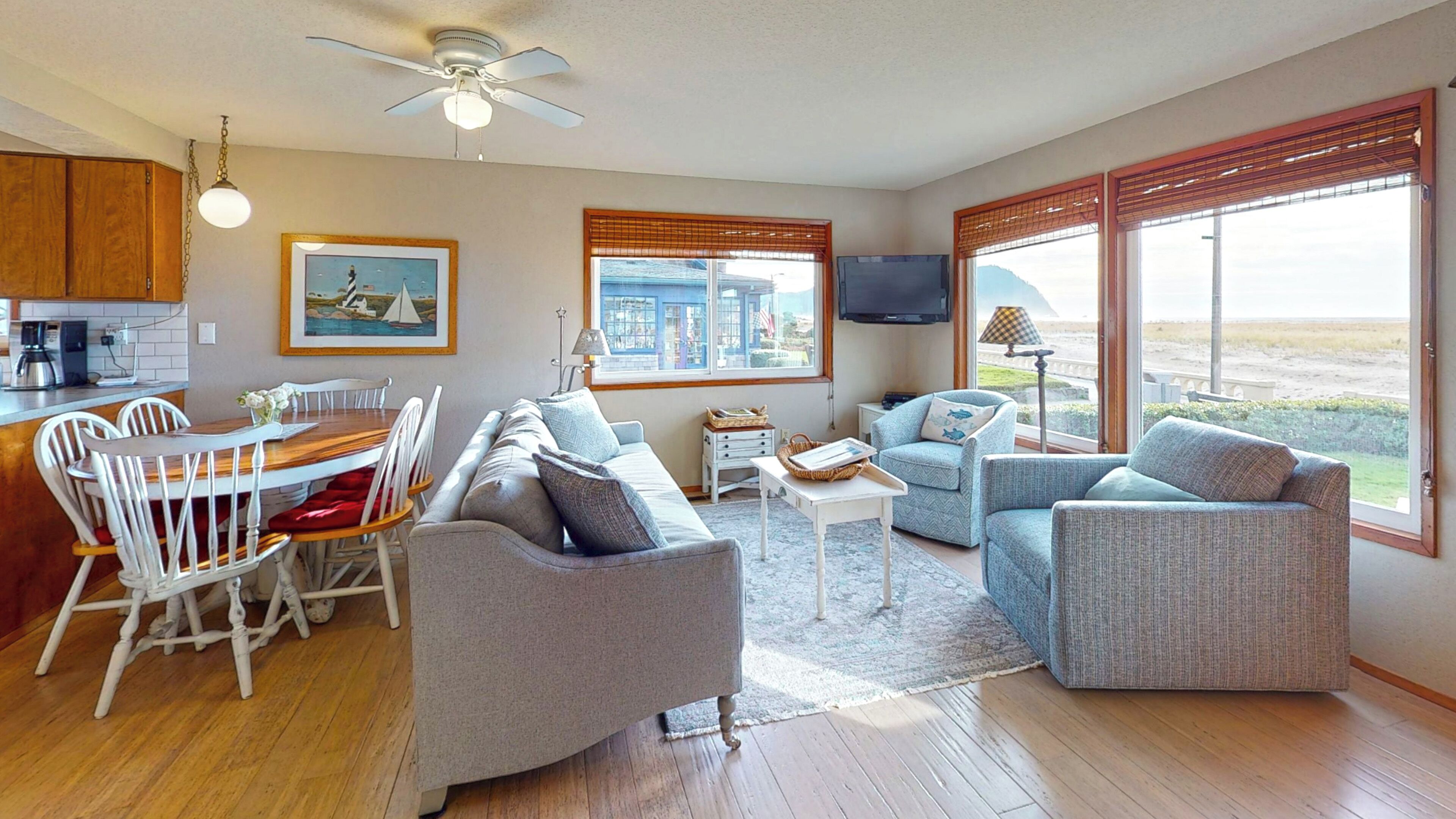 A modern living room with a dining area, large windows with blinds, a ceiling fan, and a flat-screen TV mounted on the wall.
