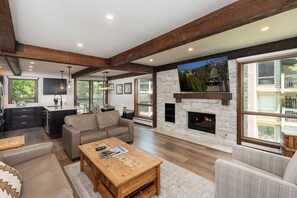 A spacious living area with a fireplace, large windows, and a kitchen in the background.