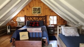 A cozy attic bedroom with a wooden headboard, a bed with a blue bedspread, two bedside tables with lamps, a small table with a fish-patterned cushion, and a window with a view of a tree. - 120Yr Old Cabin Great for couple/family, 15 min from Boone, center of 200 acres! (Boone)