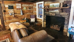 A rustic room with a fireplace, wooden walls, a leather chair, a small table, and various wall decorations. - 120Yr Old Cabin Great for couple/family, 15 min from Boone, center of 200 acres! (Boone)