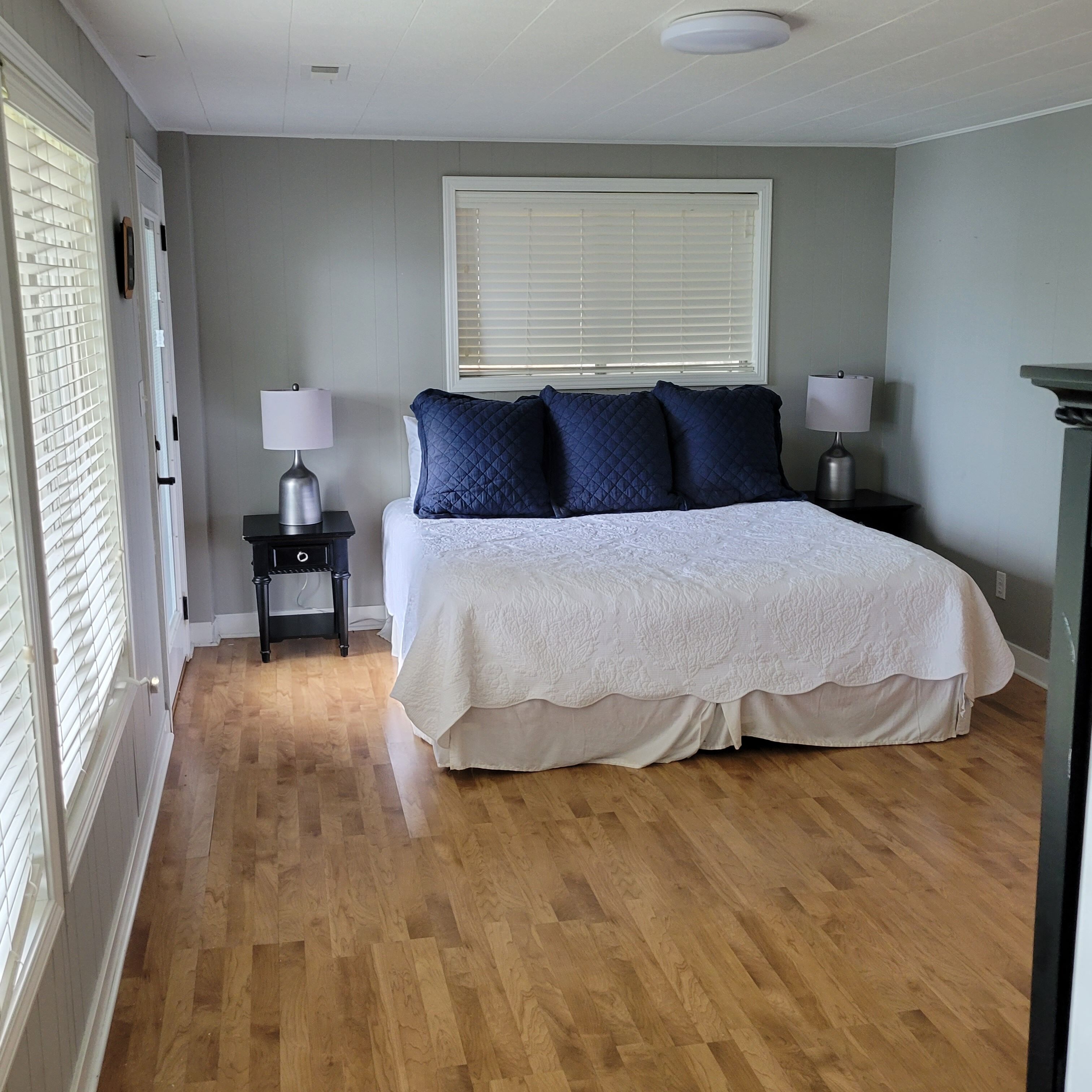 A bedroom with a large bed, two bedside tables, a window with blinds, and a wooden floor.