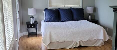 A bedroom with a large bed, two bedside tables, a window with blinds, and a wooden floor.