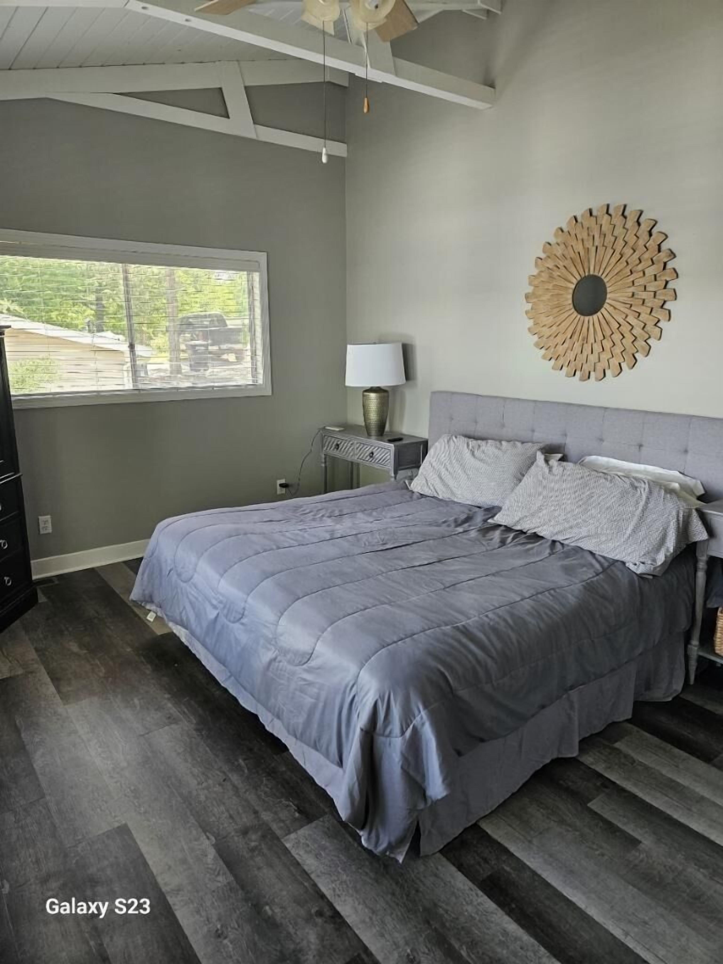 Un dormitorio con cama, mesita de noche, lámpara, un adorno en la pared y ventana con vistas al exterior.