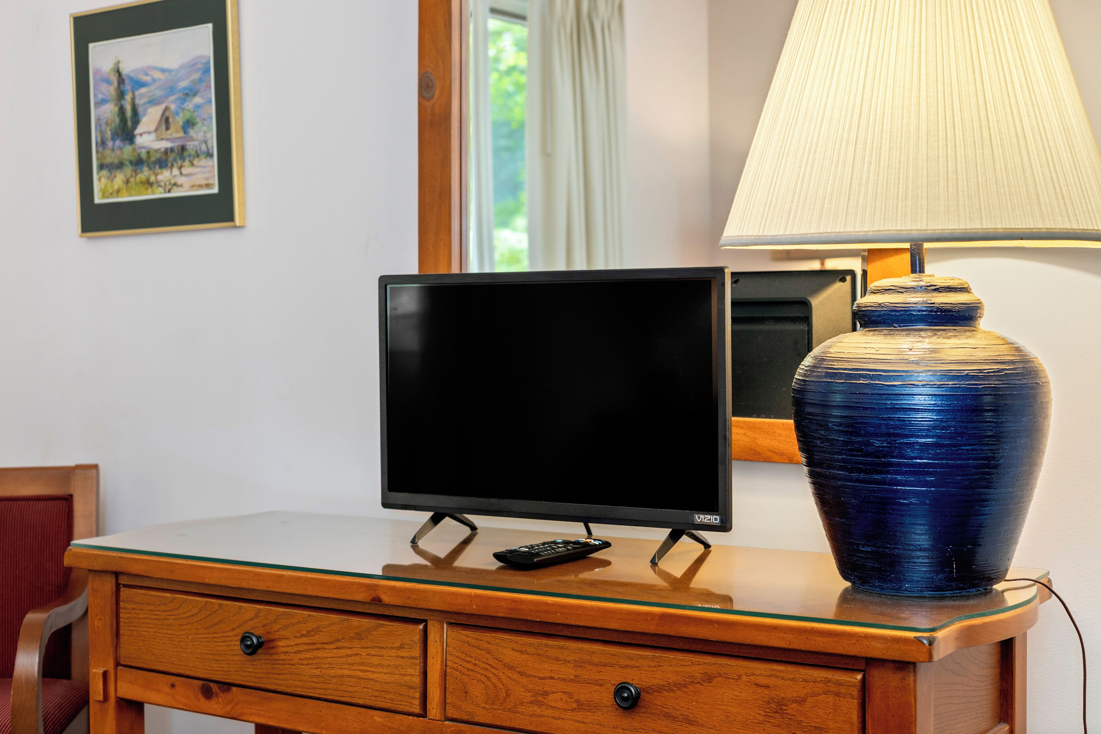 A television on a wooden dresser with a remote control, a blue vase, and a lamp.