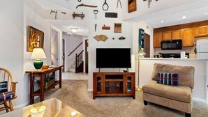 A living room with a flat-screen TV, a sofa, a dining table, and a kitchen area with wooden cabinets and a microwave.