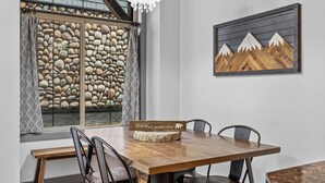 A dining area with a wooden table, four chairs, a chandelier, a stone wall, and a framed artwork. - Mountain View Chalet - Large Outdoor Pool and Hot tub - Pet Friendly (Canmore)