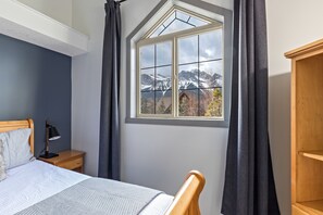 A bedroom with a bed, a nightstand, a desk, a window with a view of mountains, and a wooden wardrobe. - Mountain View Chalet - Large Outdoor Pool and Hot tub - Pet Friendly (Canmore)
