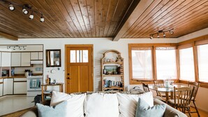 Un salon chaleureux avec un plafond en bois, une table à manger avec des chaises, une bibliothèque et un coin cuisine équipé d’une cuisinière et de placards.