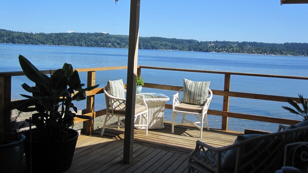 Une terrasse en bois avec deux fauteuils en osier et une petite table, donnant sur une vaste étendue d’eau bordée d’une rive boisée.