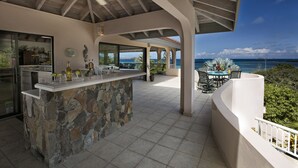 Un espacio de cocina exterior techado con una isla de piedra, un ventilador de techo y vista al océano.
